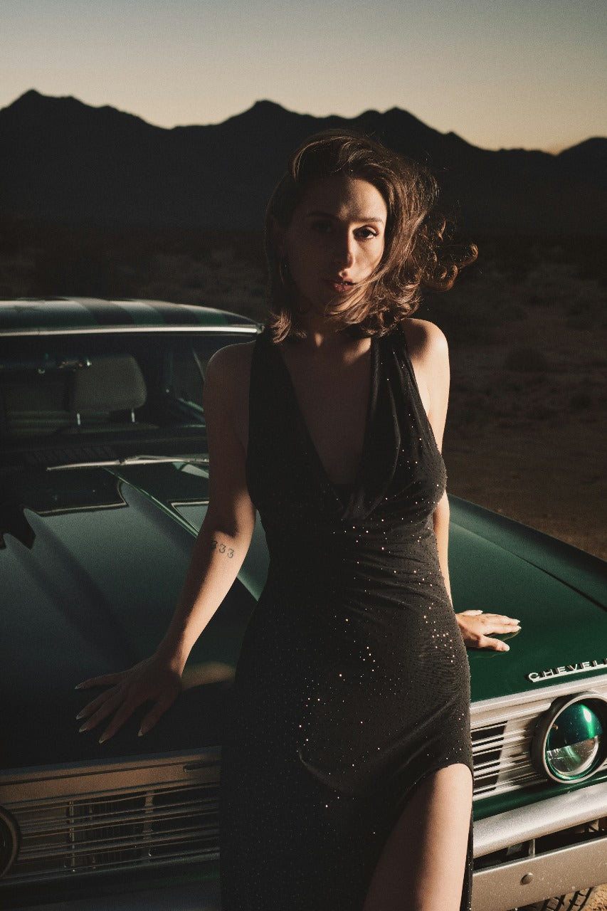 XAYWEAR black slit dress with deep neckline worn by woman leaning on vintage car at sunset