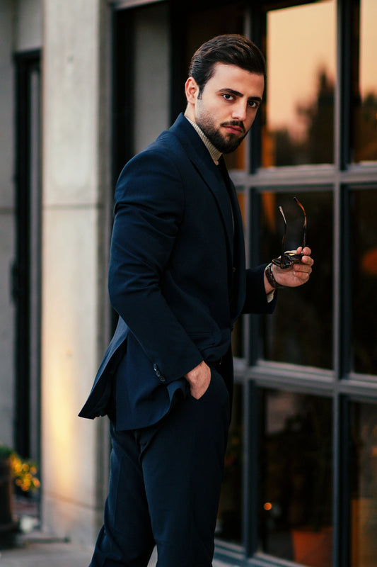 Man wearing a stylish men blue suit by XAYWEAR holding sunglasses near a window
