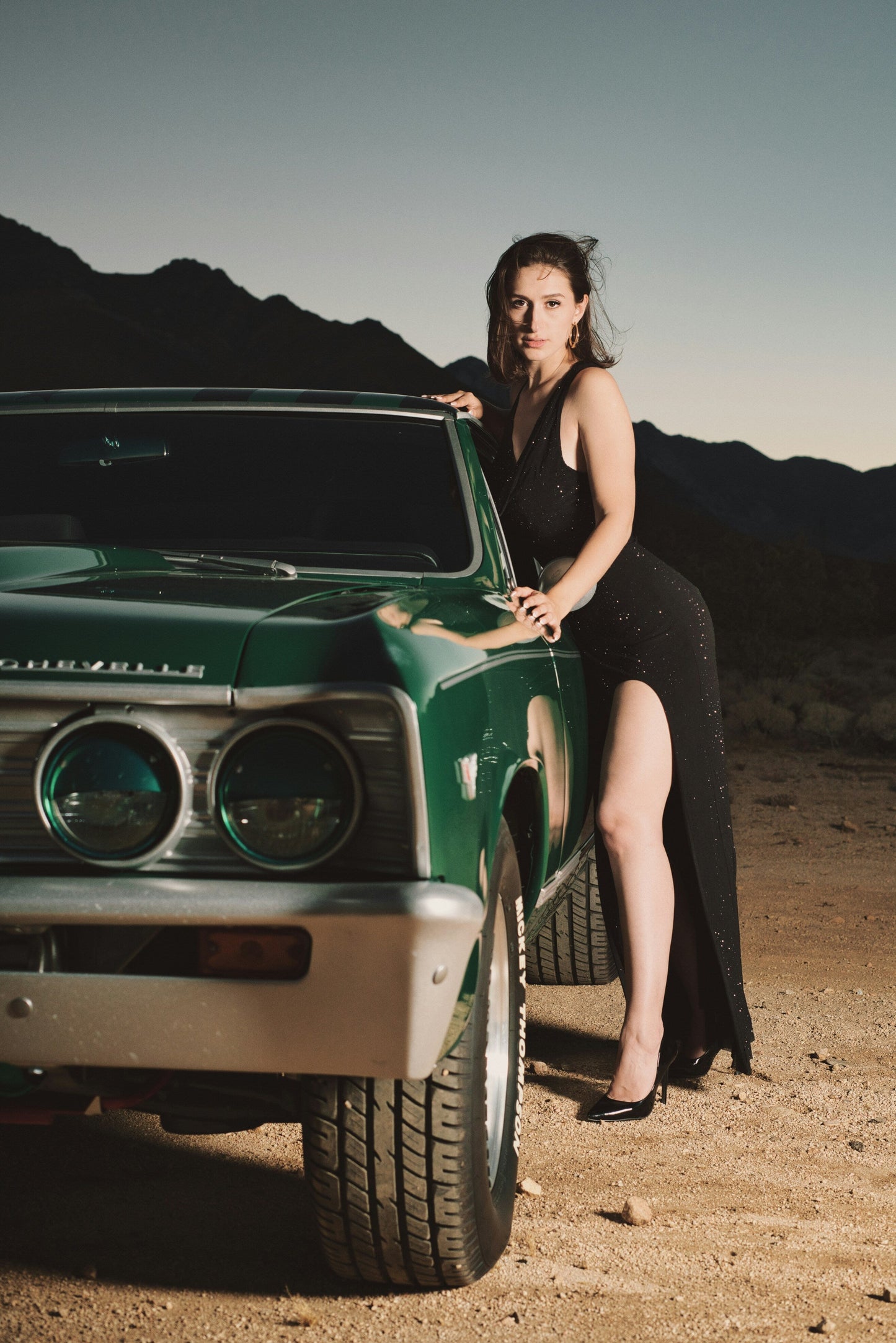 Woman wearing XAYWEAR black slit dress standing next to a vintage green car in desert at dusk