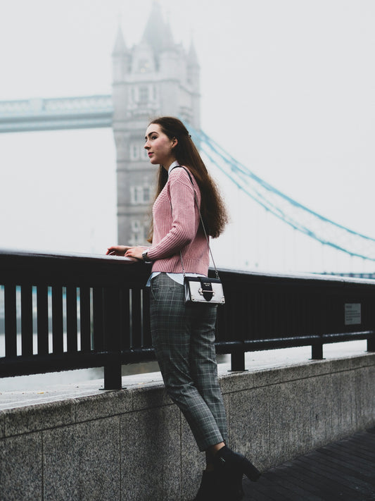 Woman wearing trench coat for women styled with plaid pants and pink sweater by the river near Tower Bridge