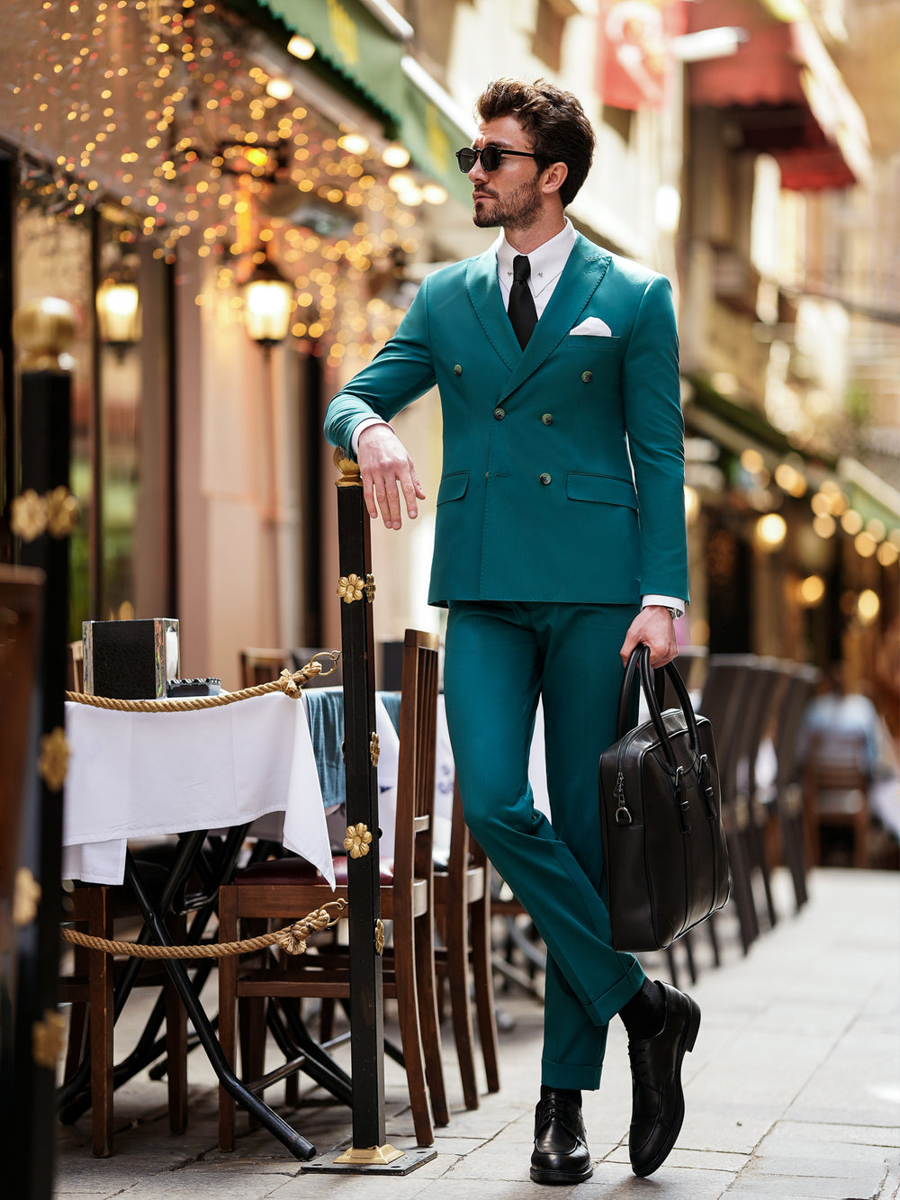 Man wearing teal suit for men by XAYWEAR standing in outdoor cafe holding black leather bag