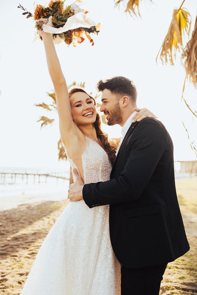 Bride wearing XAYWEAR white wedding gown holding bouquet celebrating on beach with groom