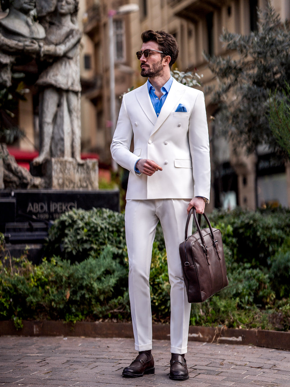 Man wearing a stylish white suit for men by XAYWEAR paired with a blue shirt and carrying a brown leather bag