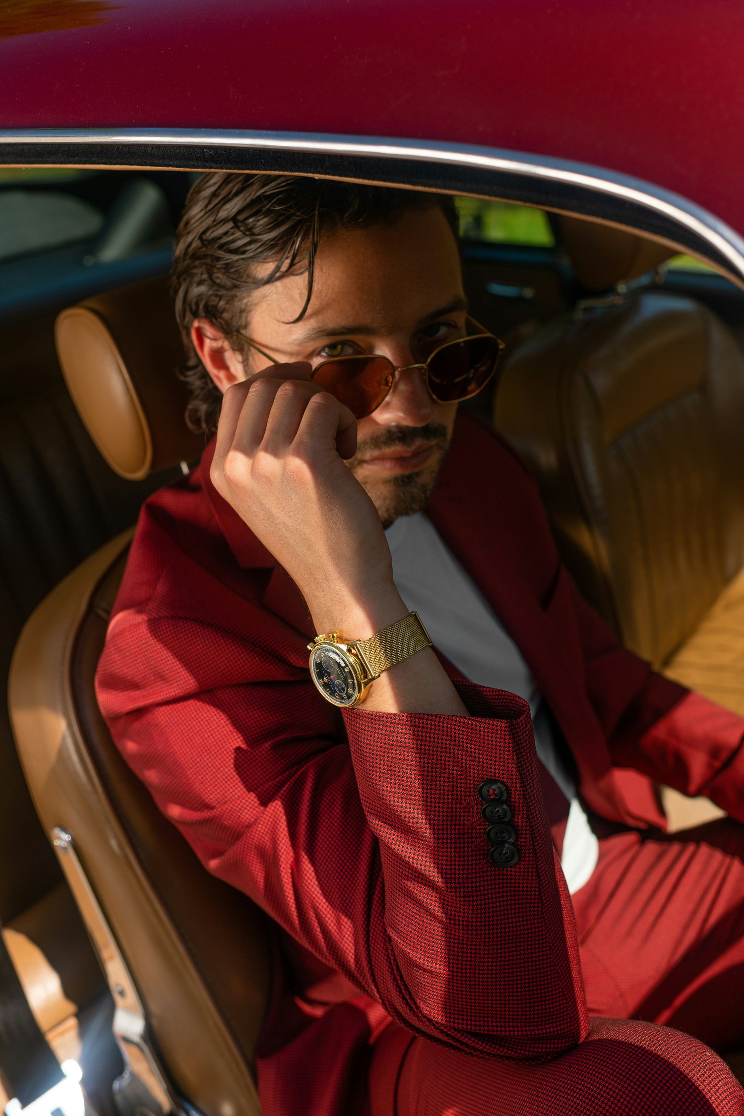 Stylish premium men wearing a red suit and gold watch sitting in a luxury car adjusting sunglasses
