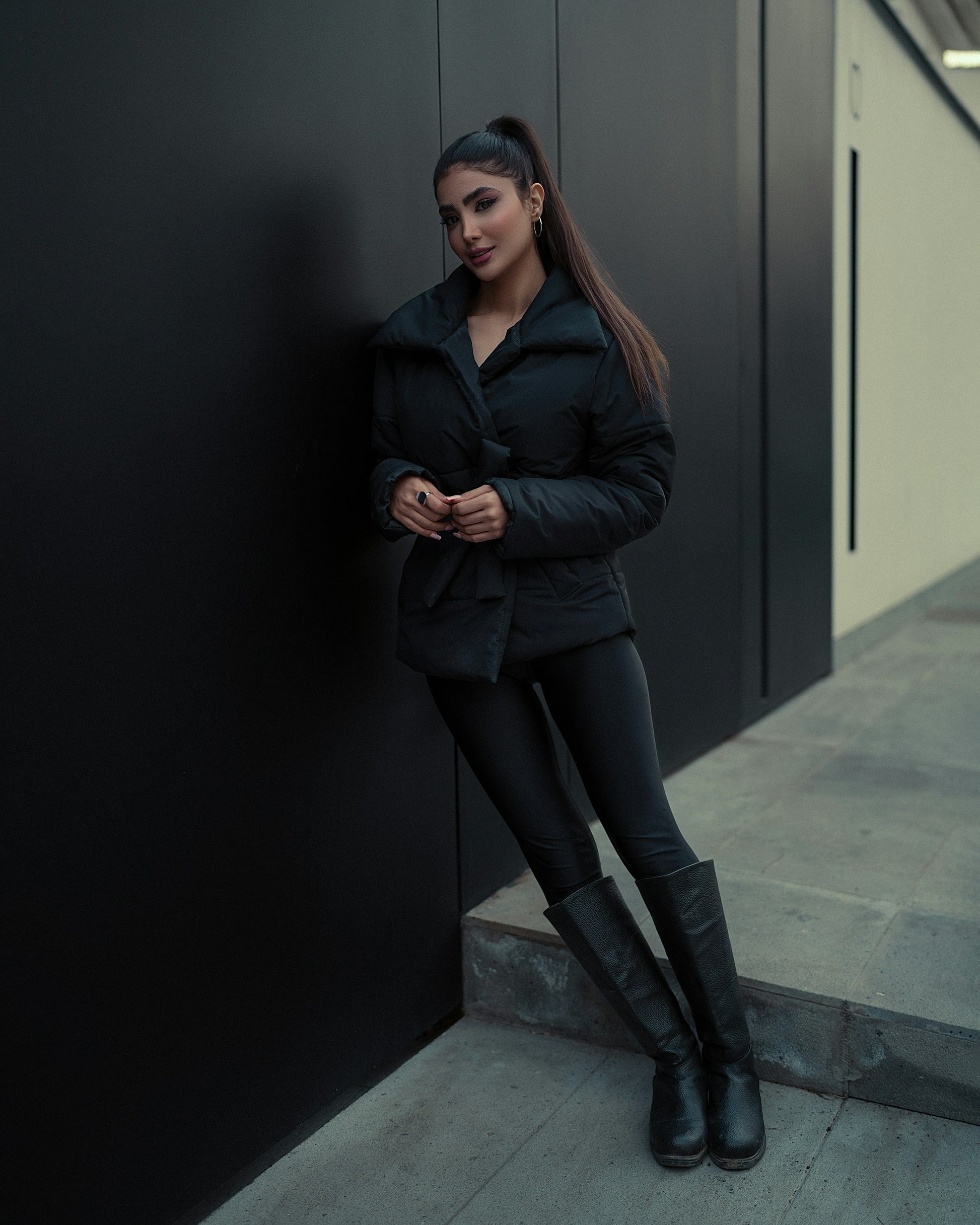 Woman wearing black puffer Jacket for women by XAYWEAR standing against a dark wall with black leggings and boots