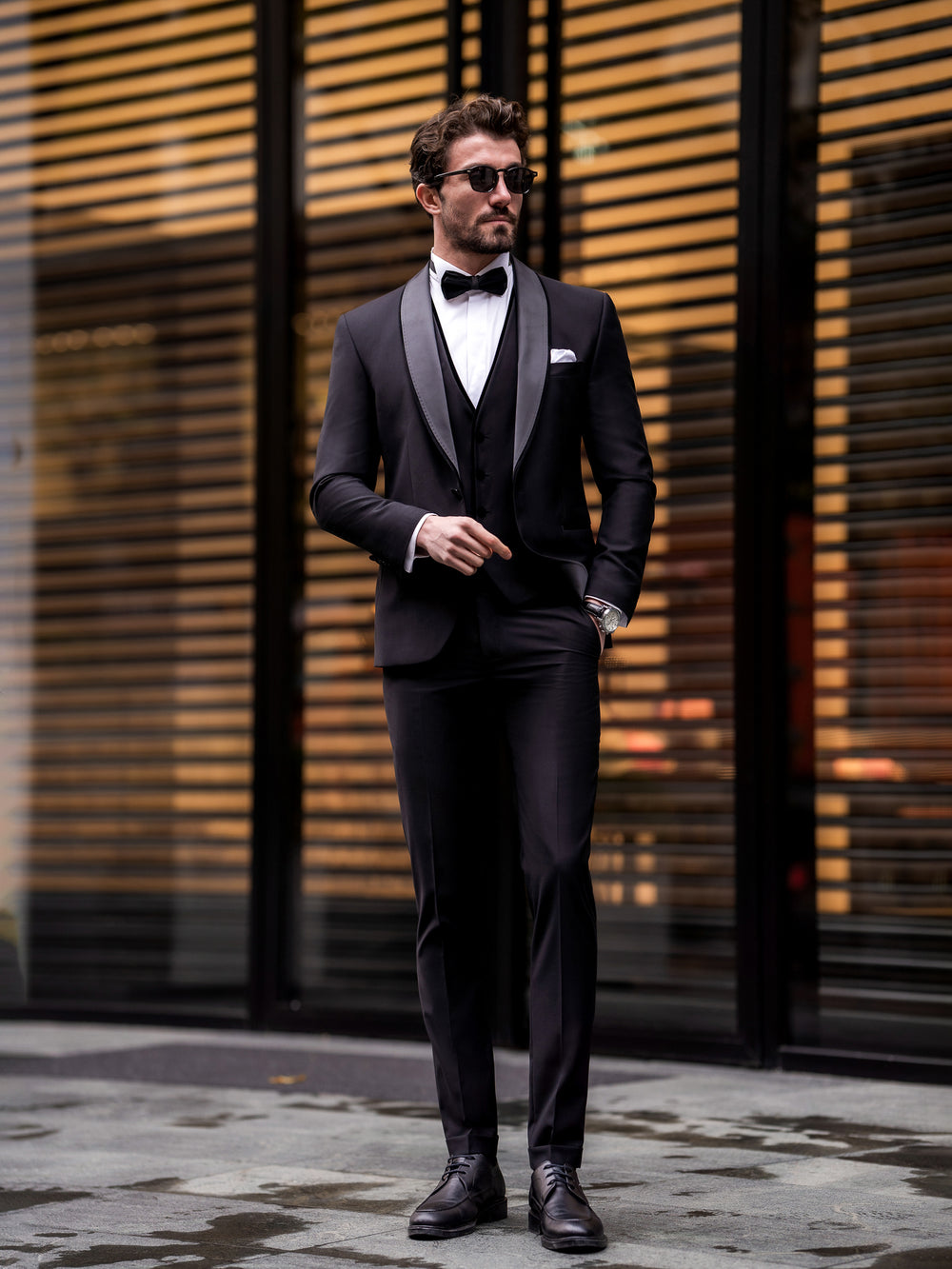Man wearing a black three piece suit for men with bow tie and sunglasses standing outdoors