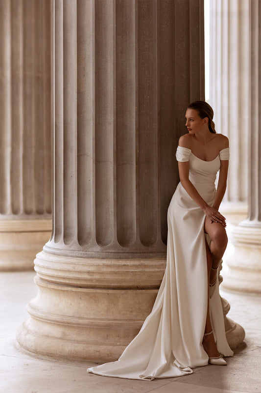 XAYWEAR elegant off-shoulder bridal gown with thigh slit worn by model sitting against classical columns