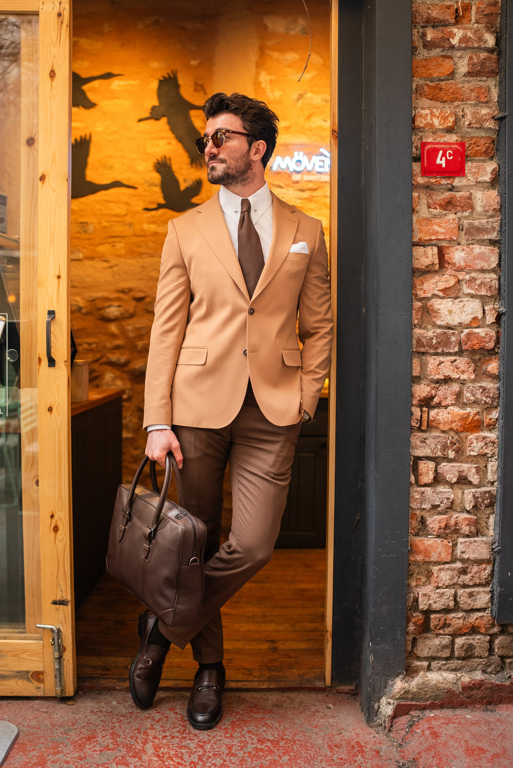 Man wearing a tan suit for men by XAYWEAR with brown tie and leather briefcase standing in doorway