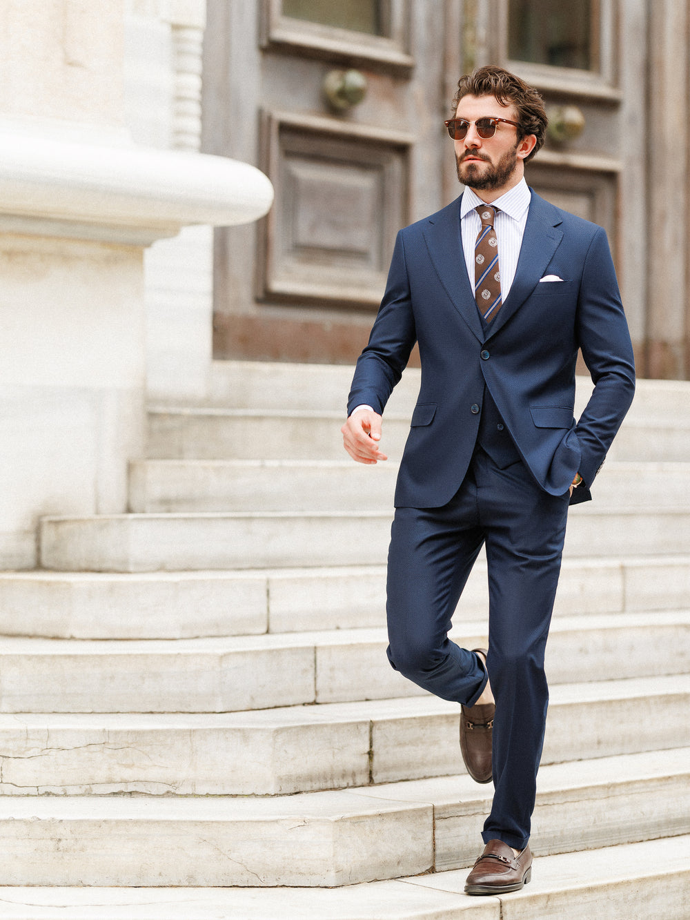 Man wearing XAYWEAR navy blue formal wedding suit with white shirt and patterned tie walking down stone stairs