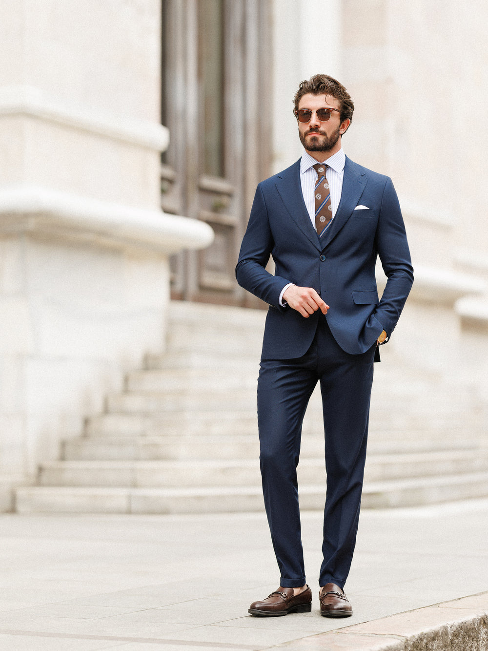 Man wearing XAYWEAR navy blue formal wedding suit with brown tie and sunglasses standing outdoors