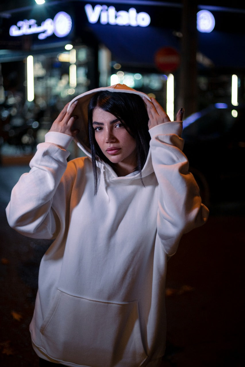 Woman wearing a white XAYWEAR hoodie with a front pocket and hood in a nighttime urban setting