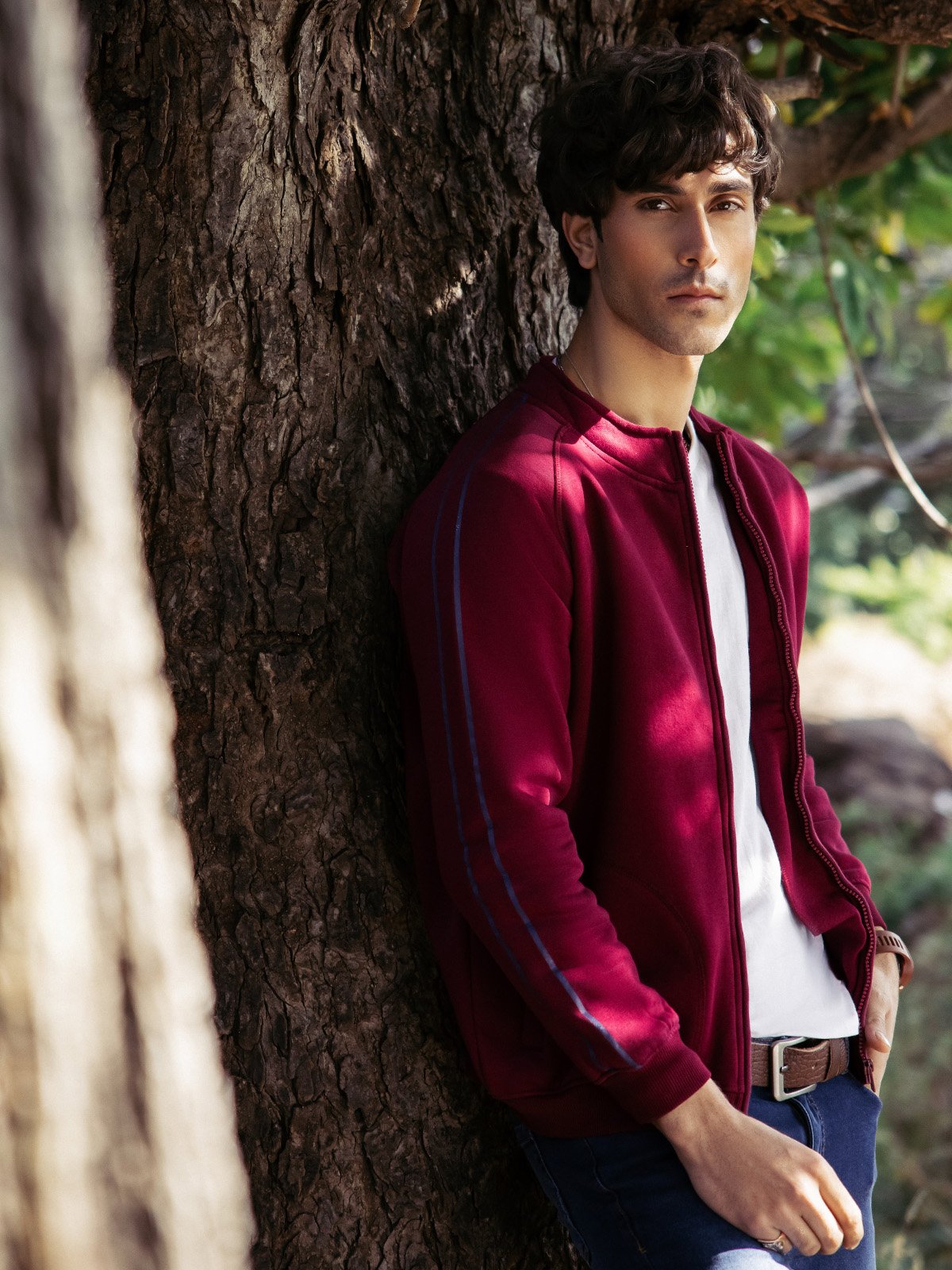 Young man wearing a maroon men casual jacket by XAYWEAR leaning against a tree outdoors