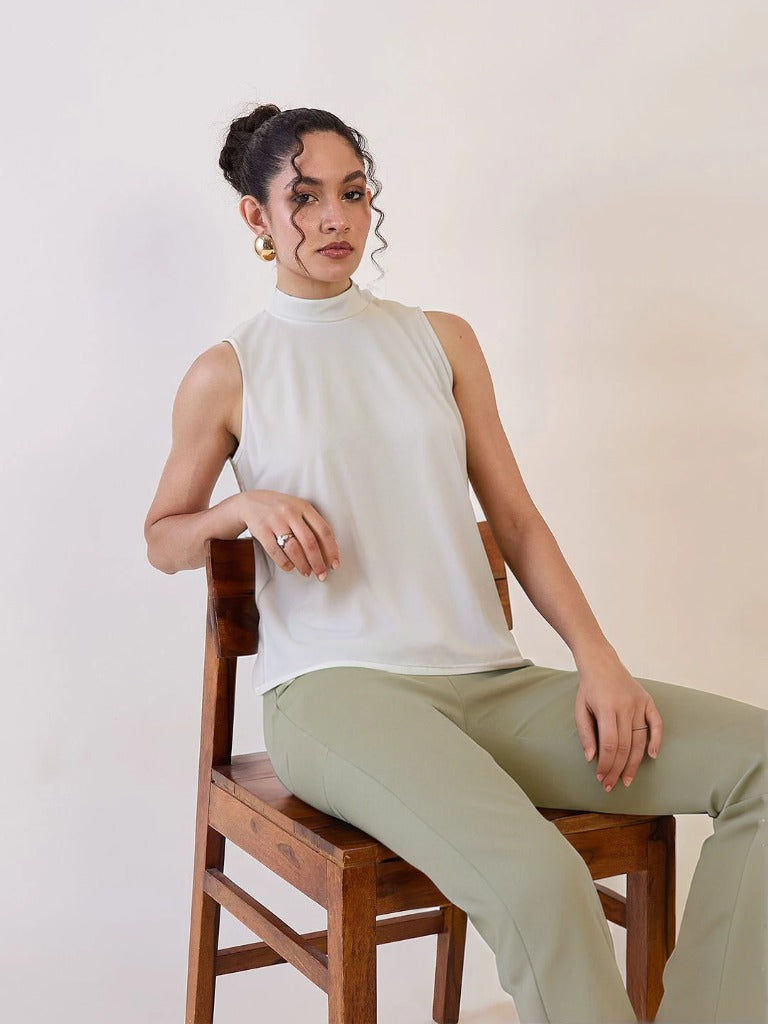 XAYWEAR white high neck sleeveless top worn by woman sitting on wooden chair with green pants