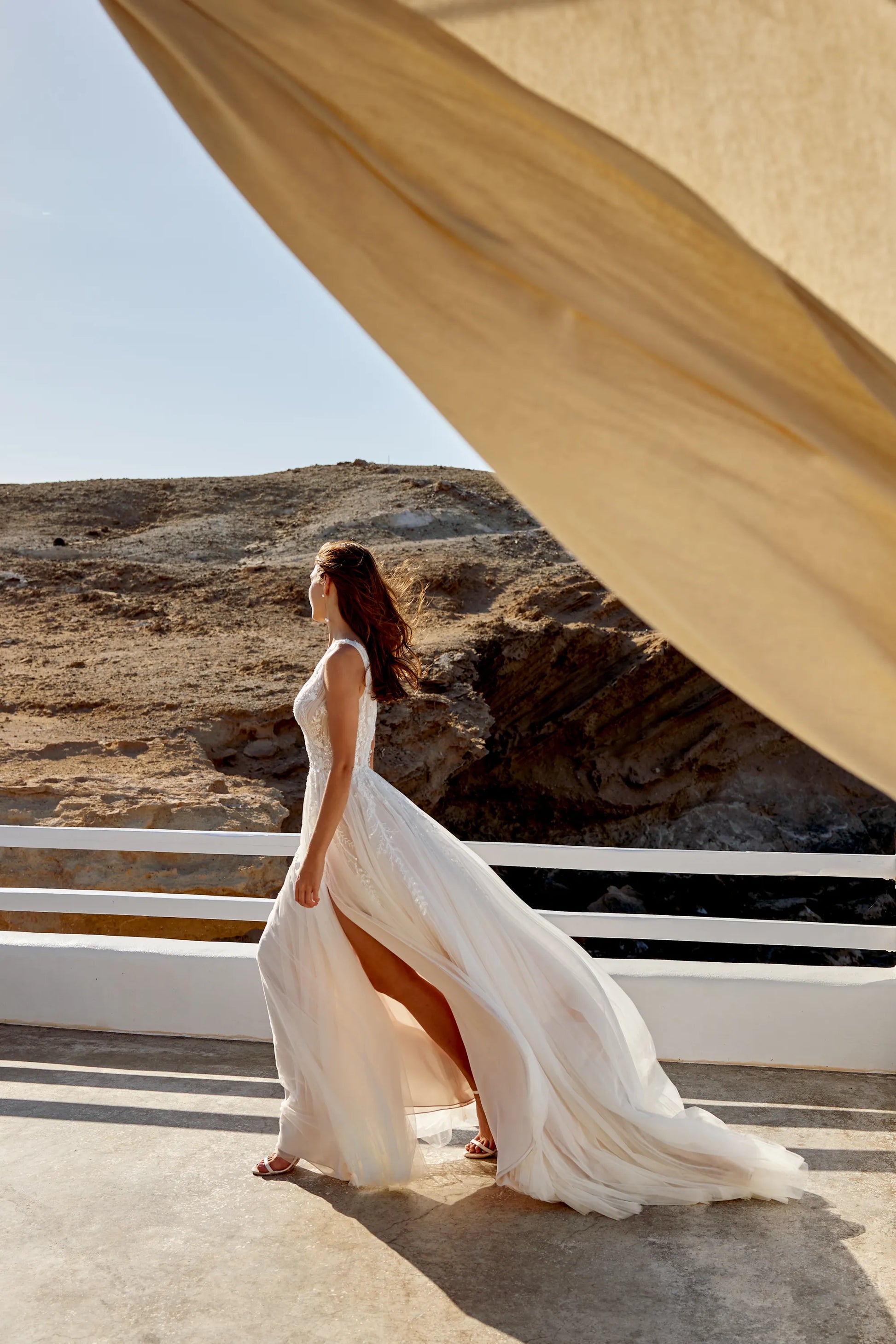 XAYWEAR wedding dress flowing in the breeze with a deep slit and lace details on a sunny outdoor terrace