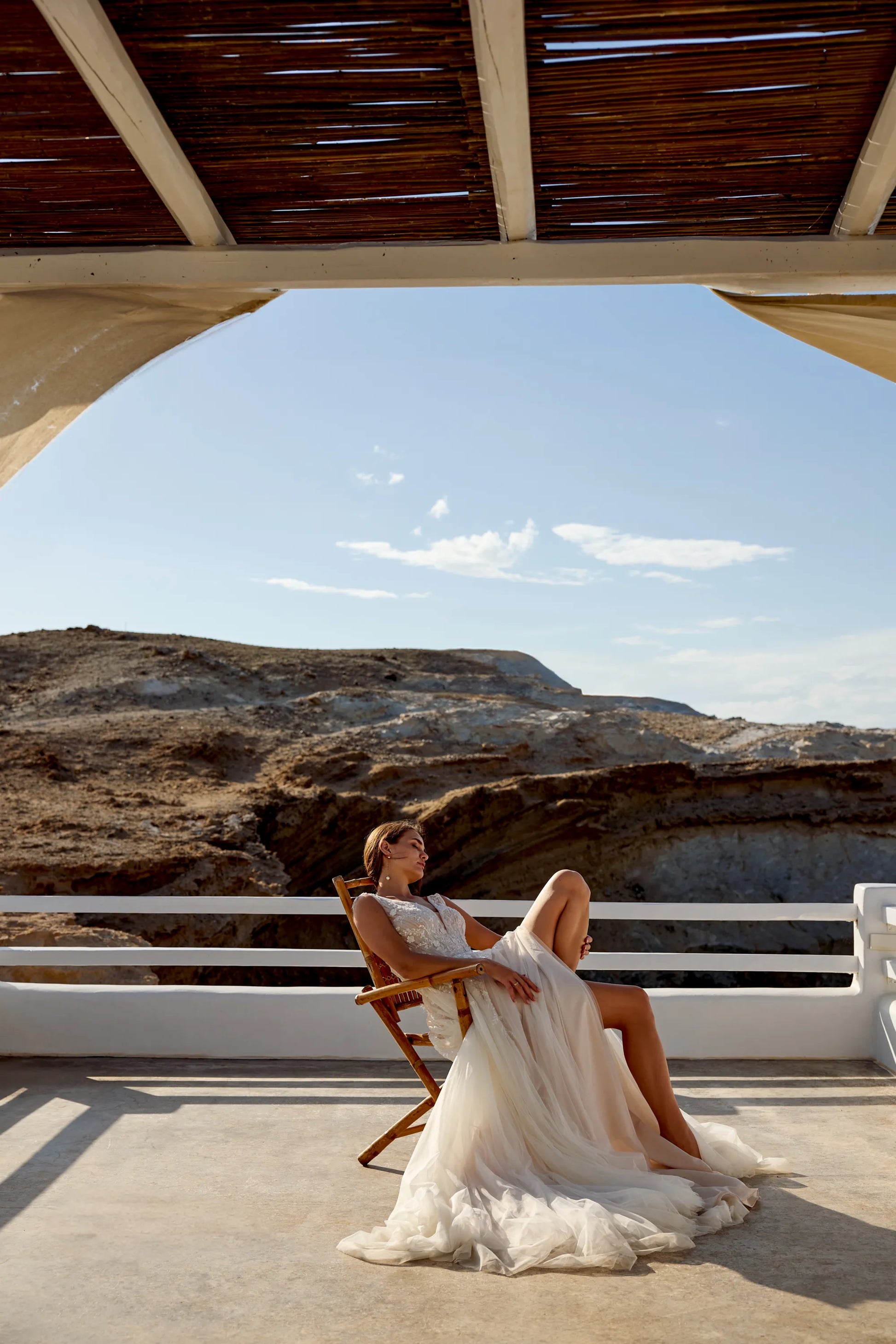 Bride wearing XAYWEAR wedding dress lounging on wooden chair with rocky landscape background