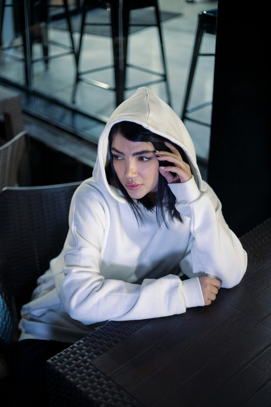 Woman wearing a white XAYWEAR hoodie sitting at a table with the hood up indoors